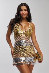 Woman with long dark hair wearing the SHAKE IT UP SEQUIN MINI DRESS IN GOLD MULTI, holding a shiny gold clutch against a plain light gray background.